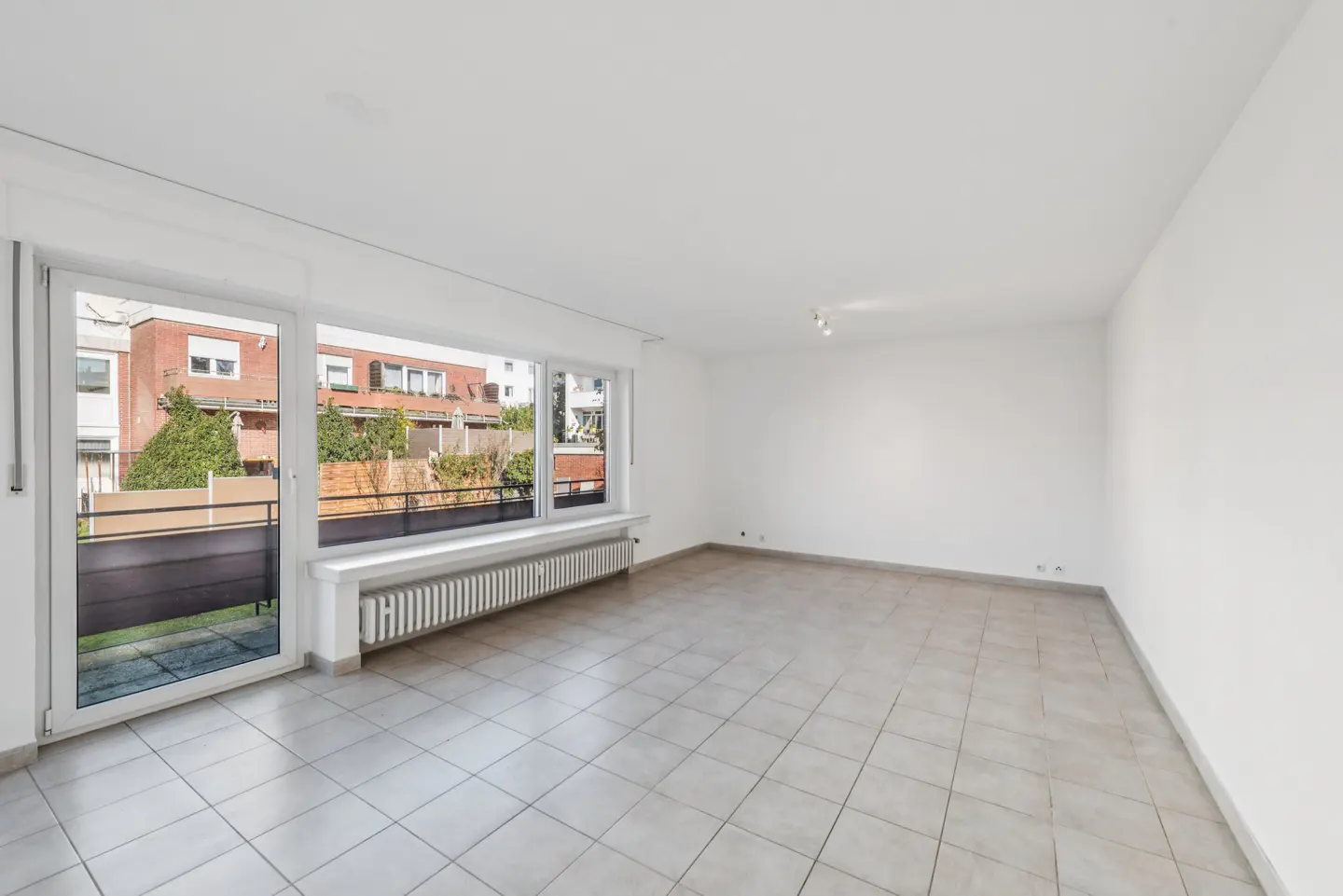 Bright, empty room with white walls and tiled floor. Large windows overlook a balcony and neighboring buildings. A radiator sits beneath the windows.
