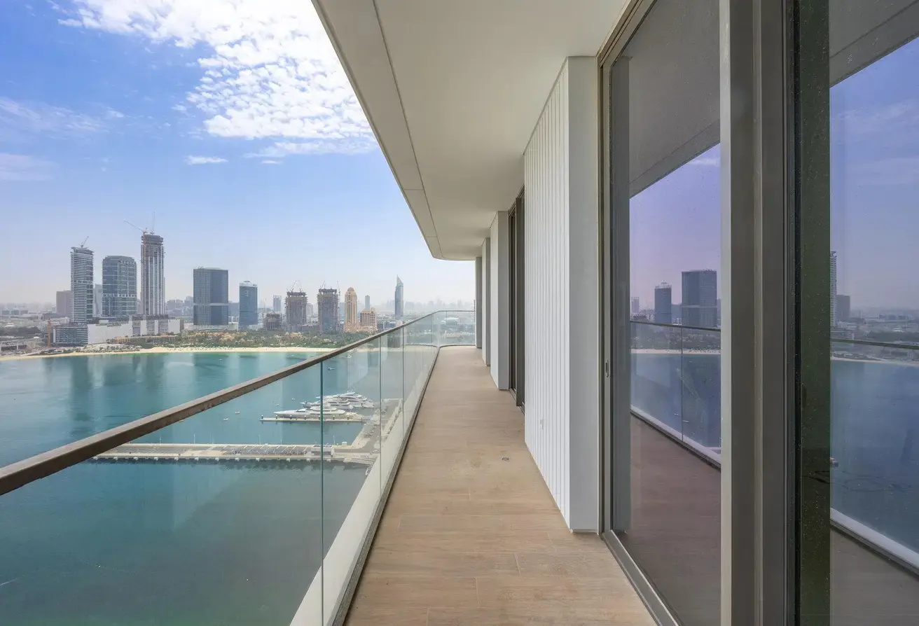 Balcony view of Dubai skyline. Glass railing overlooks the water and city buildings under a blue sky. Wood-look flooring.