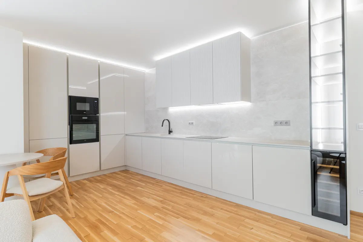 Bright, modern kitchen with white cabinets, wood floors, black appliances, and a wine fridge. A table and chairs are visible.