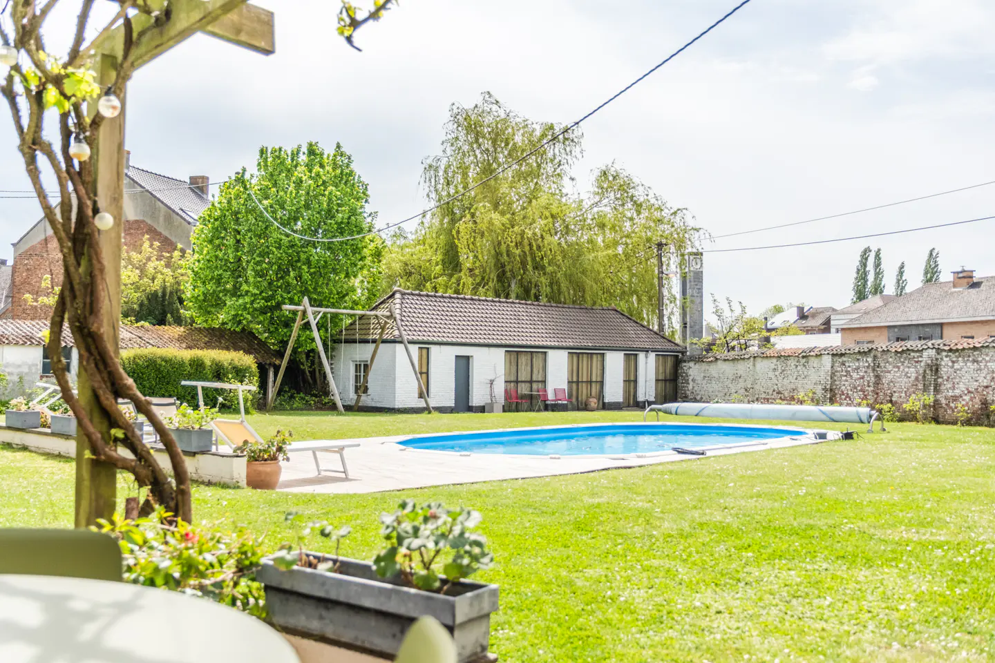 A backyard with a blue pool, green grass, a white building, and a swing set.