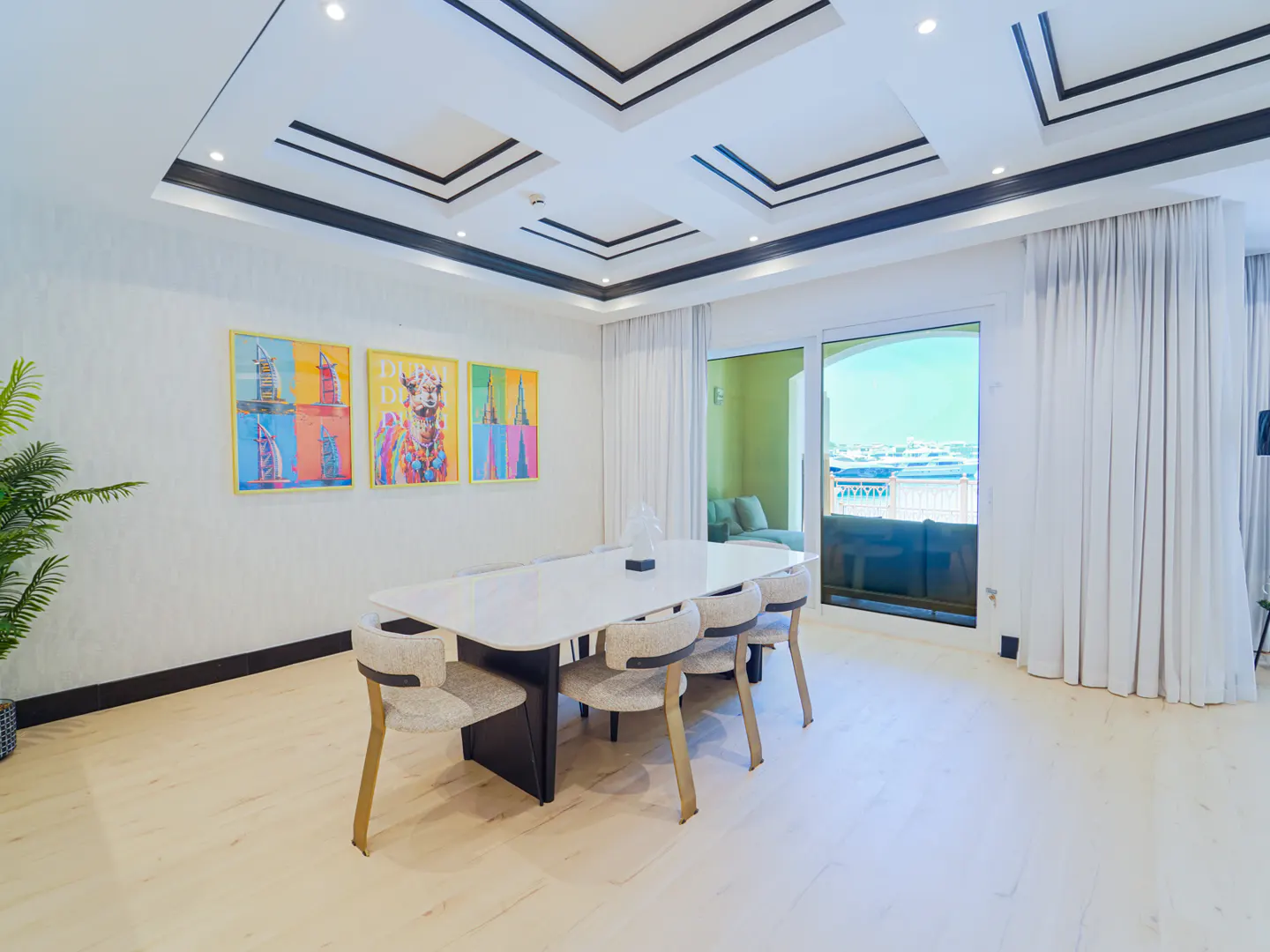Bright dining room with a white table, six chairs, and colorful artwork on the wall. A balcony offers a view of the water.