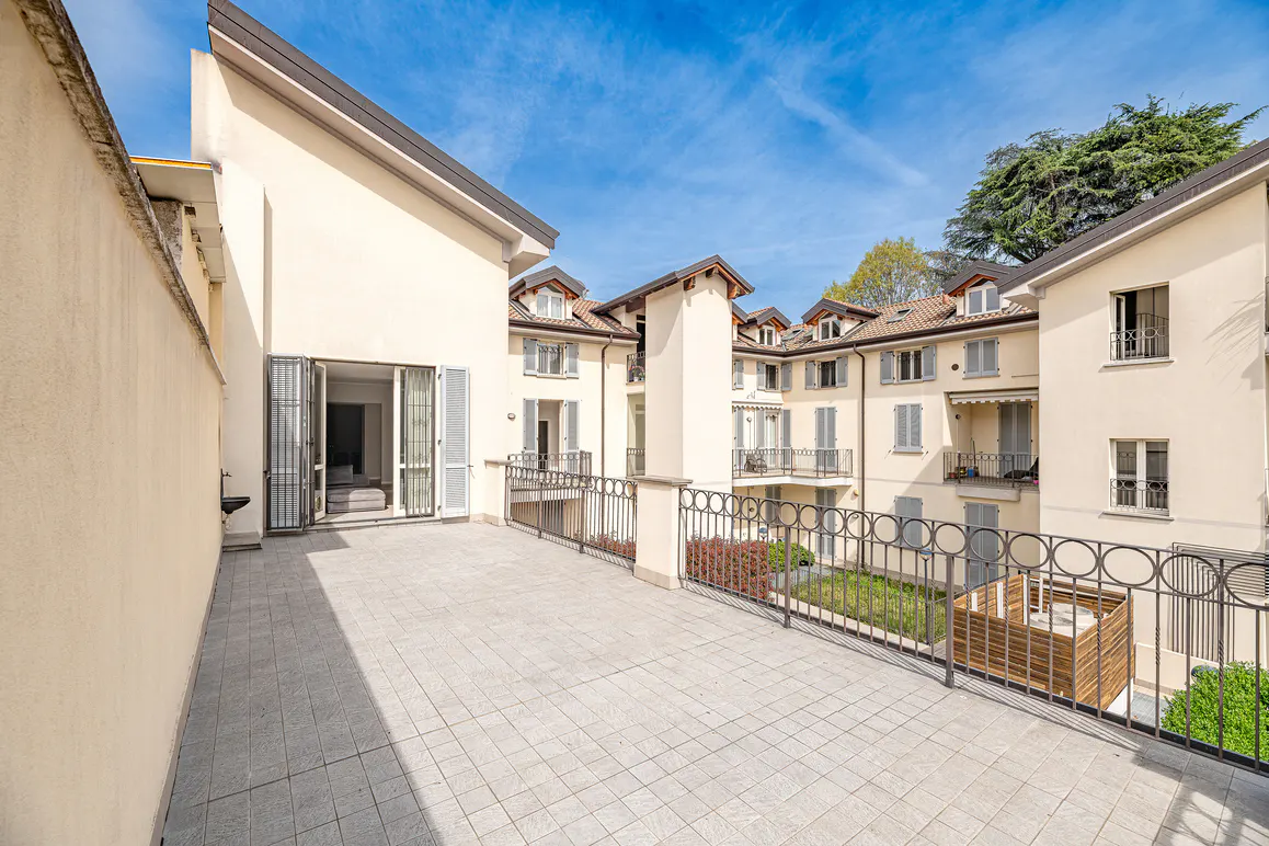 A large, tiled patio with a black metal railing overlooks a courtyard with a small lawn and garden. Cream-colored buildings surround the courtyard under a blue sky.