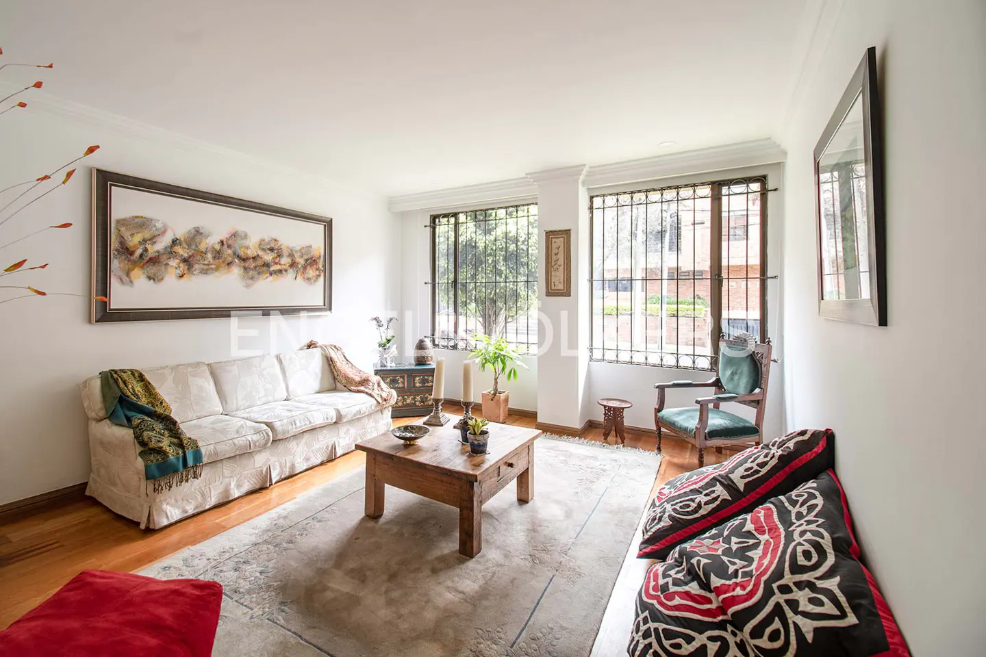 Living room with white walls, wood floors, and a large area rug. A white sofa, wooden coffee table, and green armchair furnish the space. Windows with bars look out to greenery.