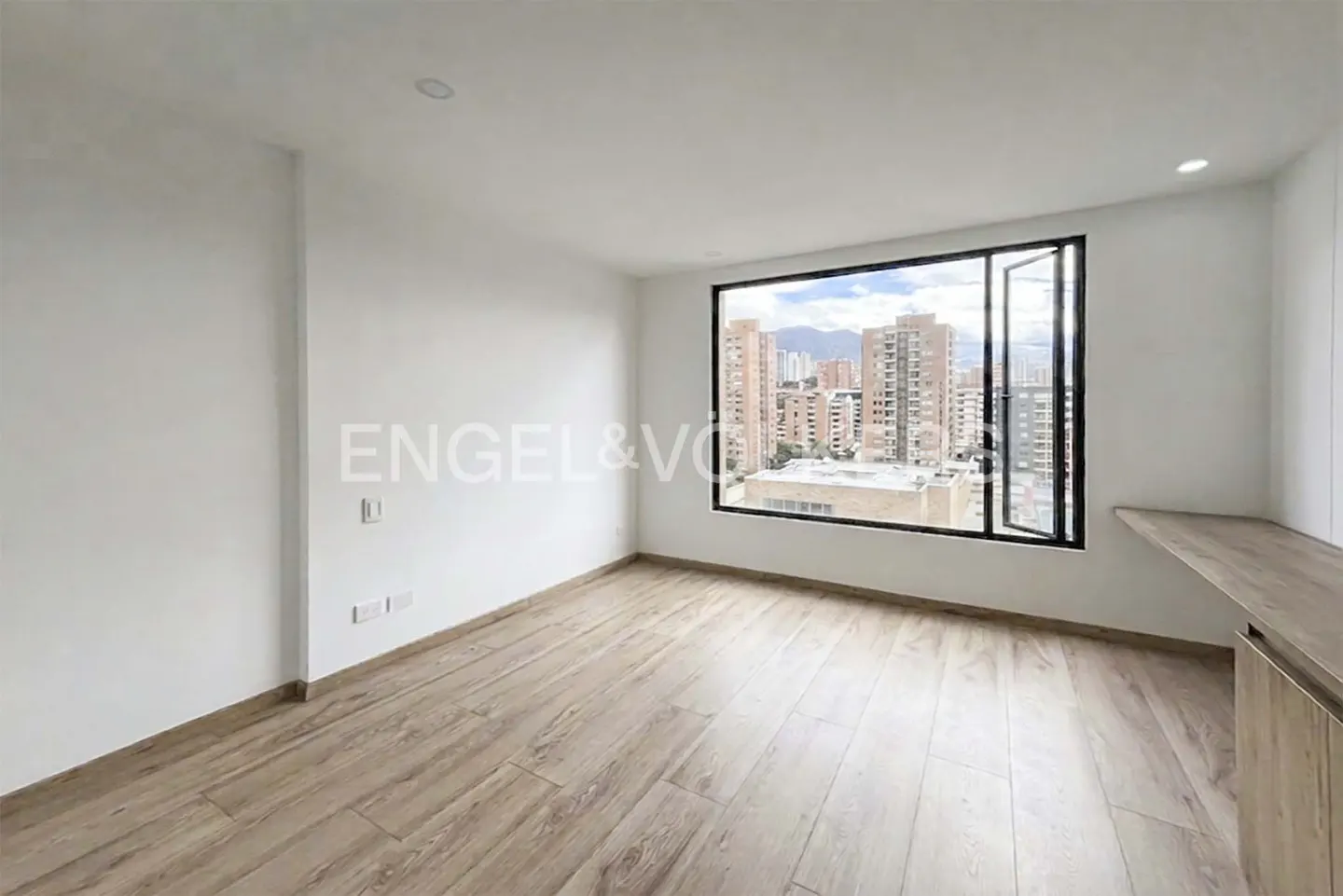 Bright, empty room with wood floors and a large window. City buildings are visible through the window. A built-in desk is on the right.