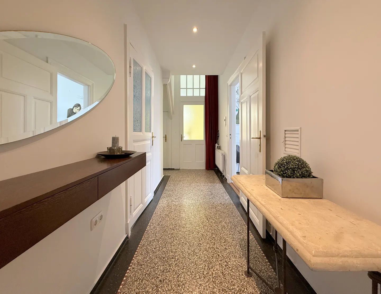 Bright hallway with terrazzo flooring, white walls, a floating dark wood shelf, and a stone console table with a plant.