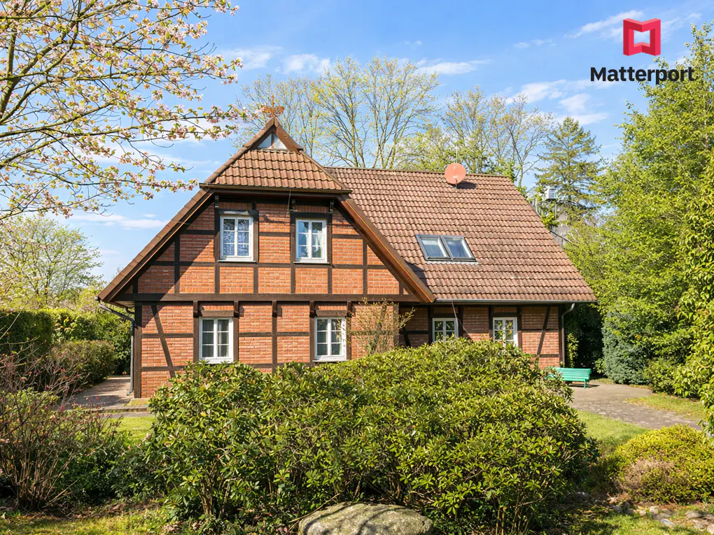 Two-story brick house with a brown-tiled roof and white-framed windows, surrounded by green bushes and trees.