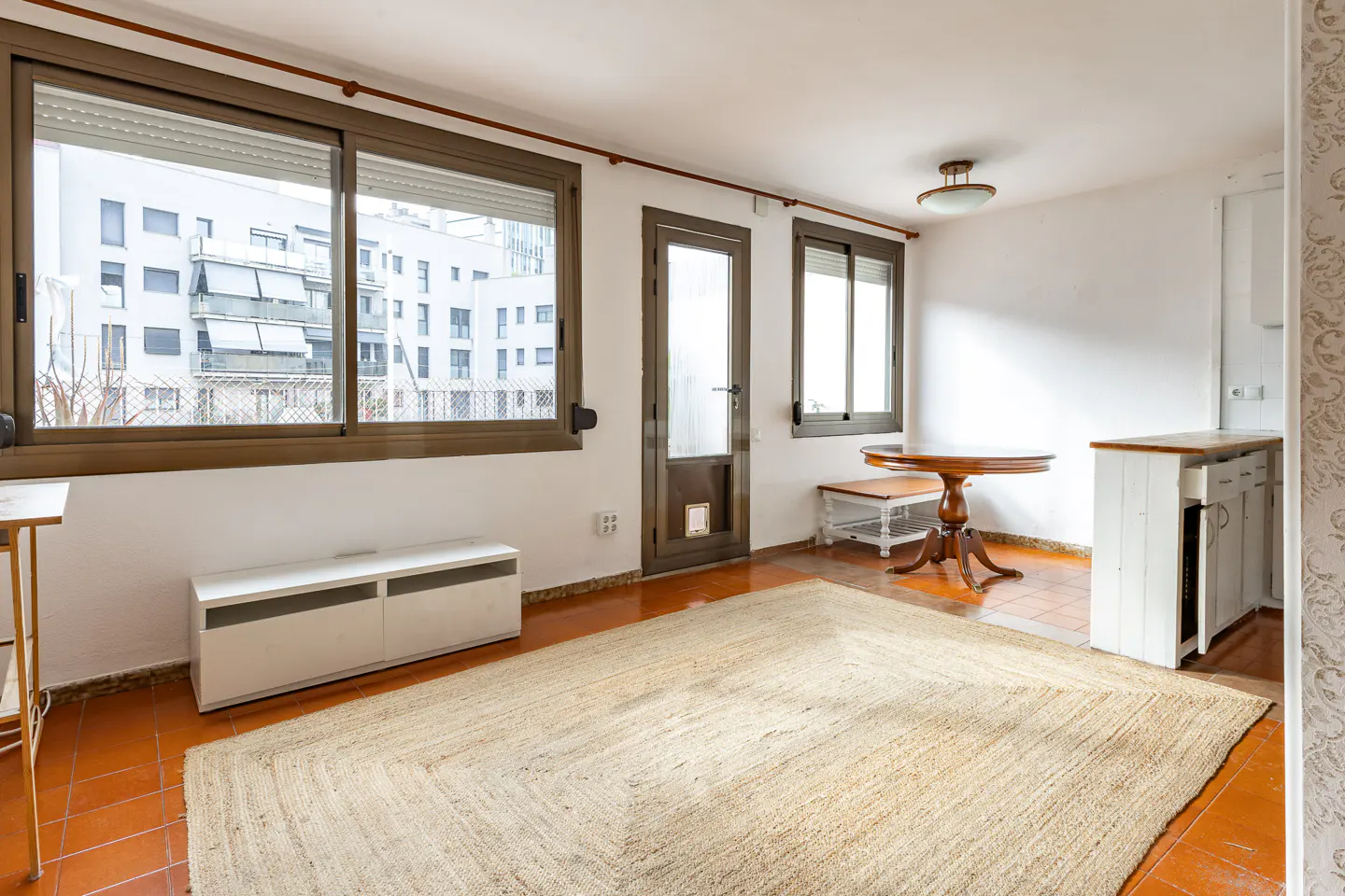 Bright living room with a large window, a round wooden table, and a beige rug on terracotta tile floor.