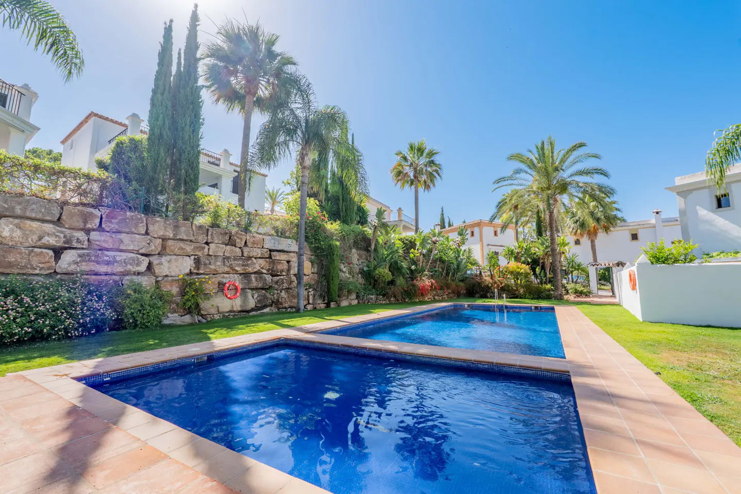 Two blue swimming pools set in a sunny garden with palm trees, a stone retaining wall, and white villas in the background.