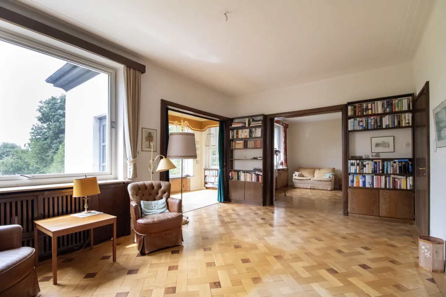 Spacious living room with parquet floors, leather armchairs, floor lamps, and built-in bookshelves filled with books.