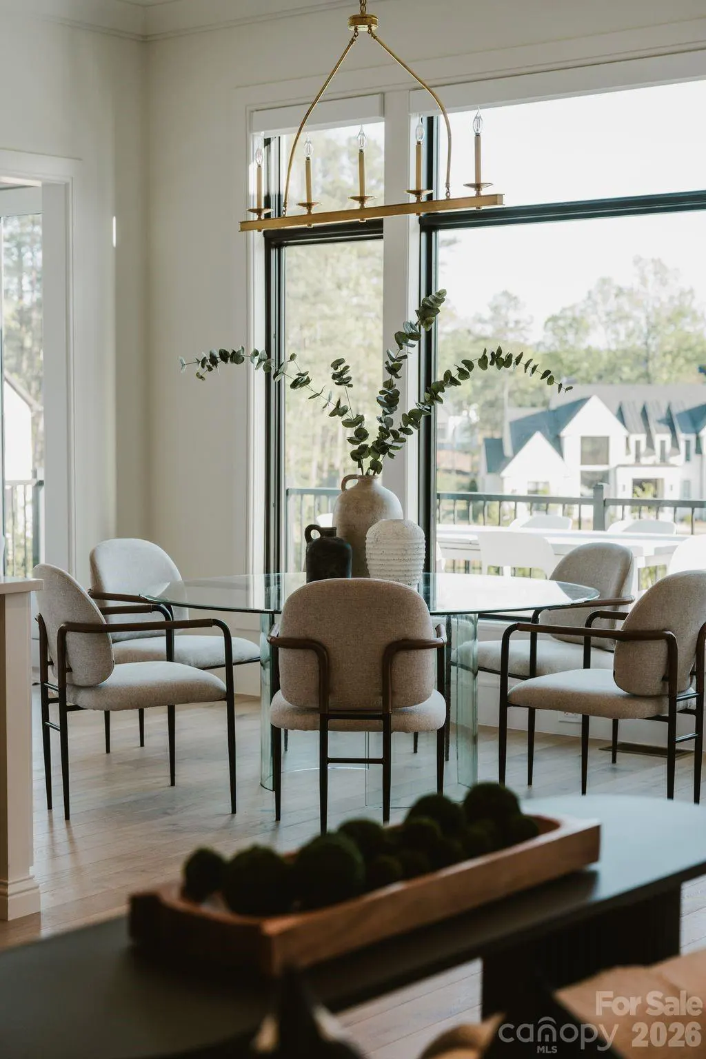 Modern dining room with a glass table, cream-colored chairs, a gold chandelier, and a large window overlooking a house.