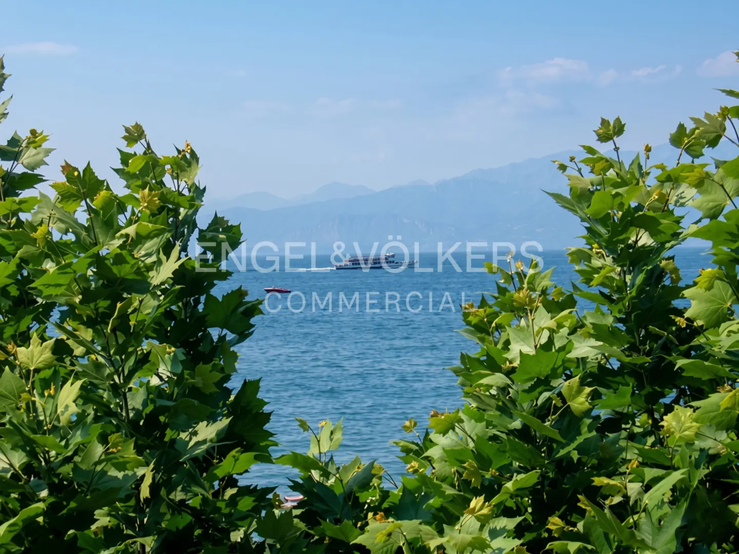 View of a blue lake with boats, framed by green trees, under a clear sky with distant mountains.