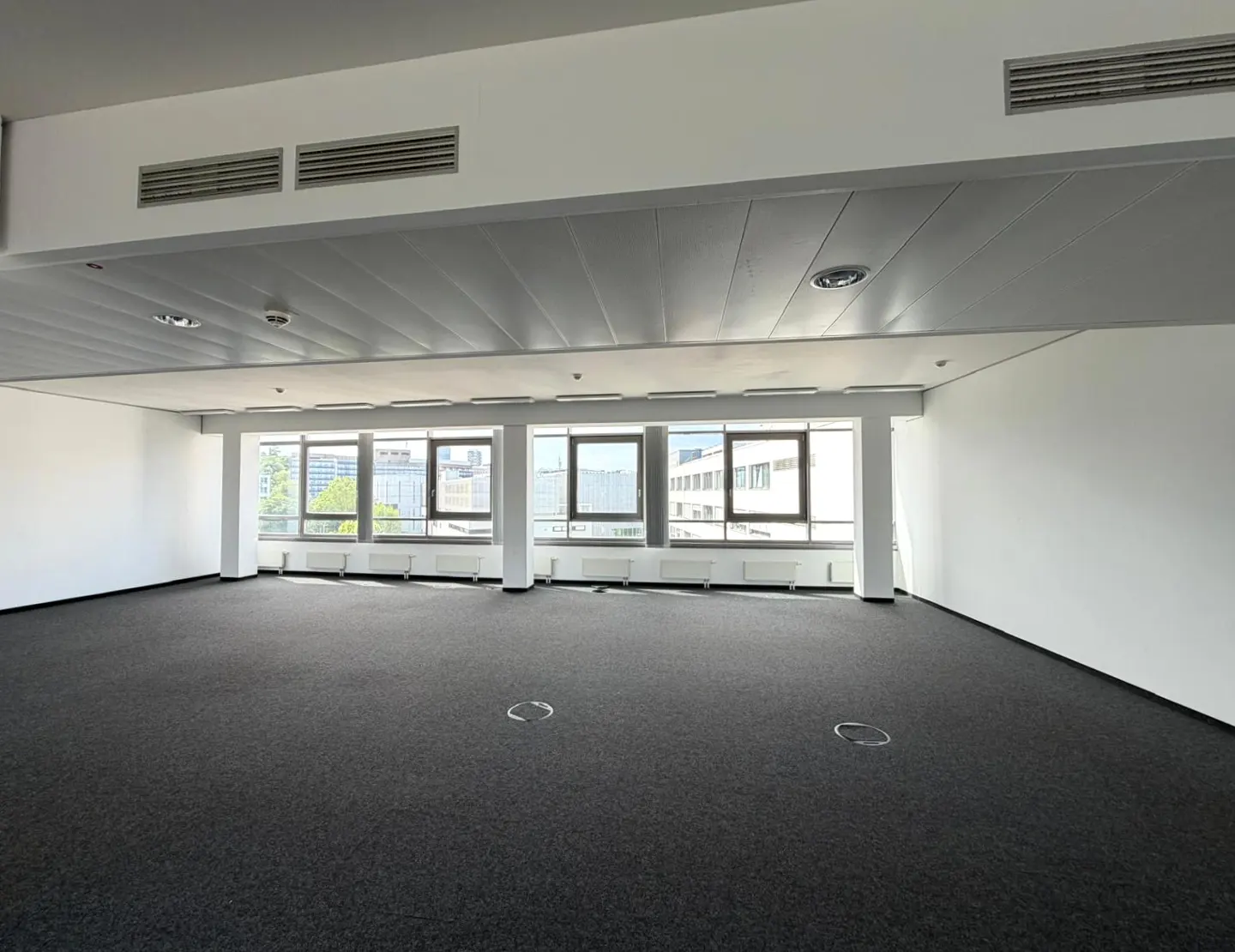 Empty office space with gray carpet, white walls, and large windows showing city buildings.