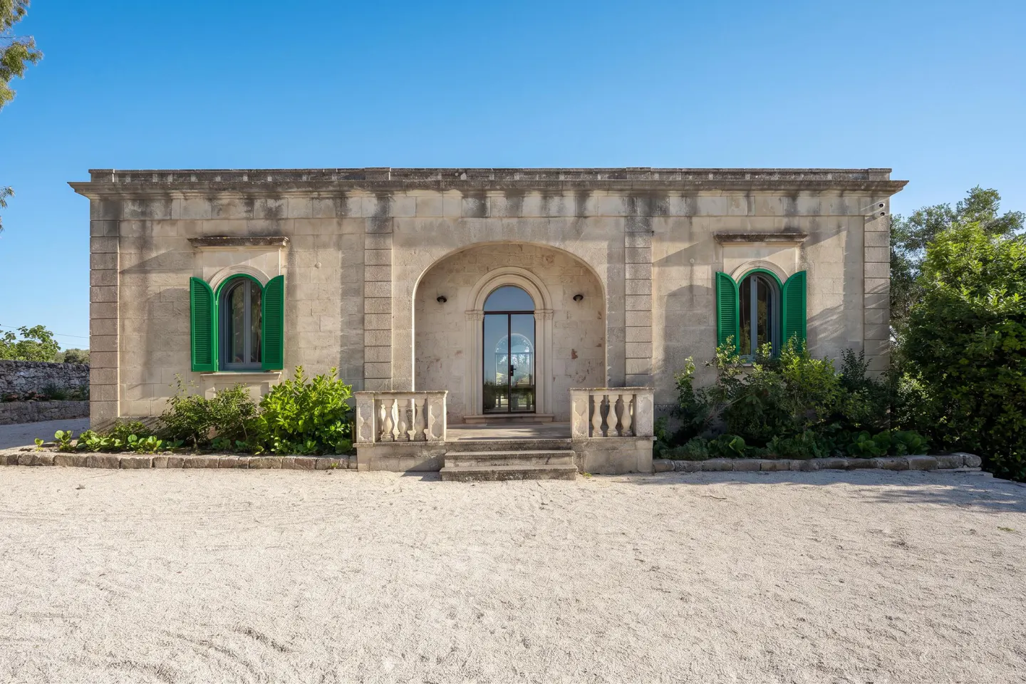 Single-story stone villa with arched doorway, green shutters, and a gravel driveway under a clear blue sky.