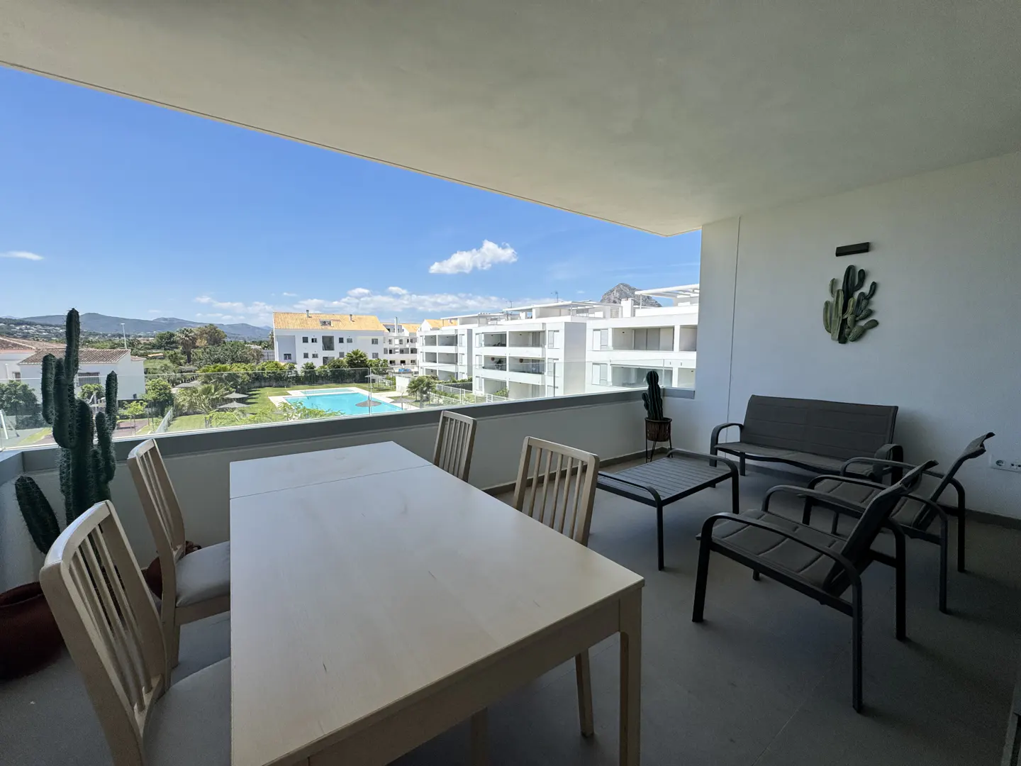 Balcony with a table, chairs, and a sofa overlooking a pool and buildings on a sunny day.