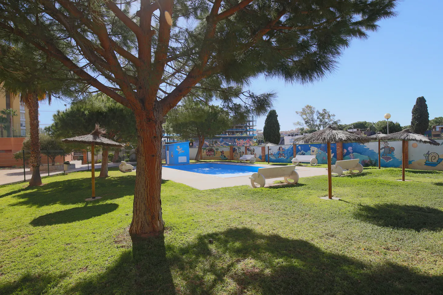 Outdoor pool area with blue water, surrounded by green grass, benches, and straw umbrellas. A mural decorates the back wall.