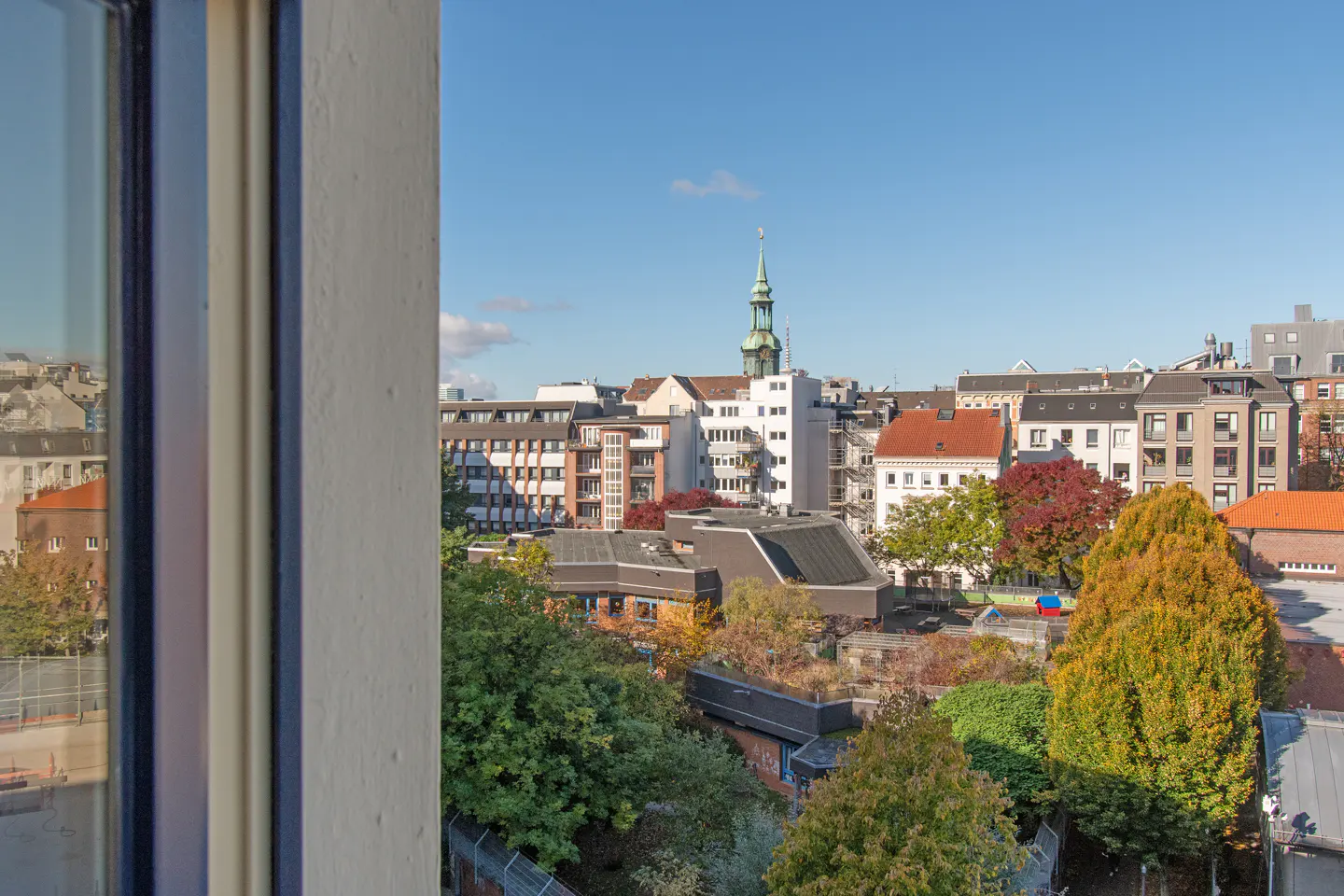 View from a window overlooking a European city with buildings, trees, and a church steeple under a blue sky.