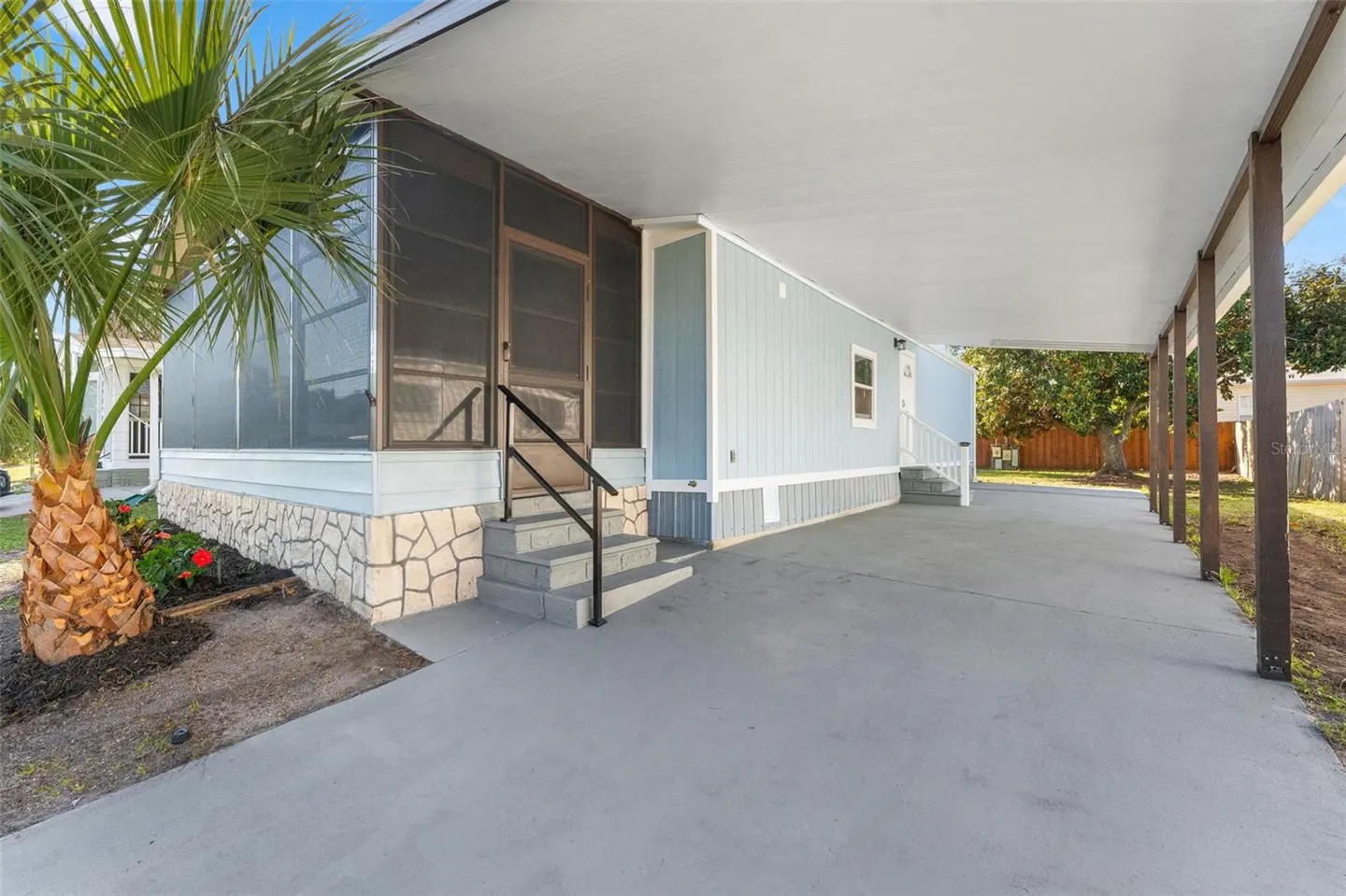 Exterior view of a light blue mobile home with a carport, a screened-in porch, and a palm tree in the front yard.