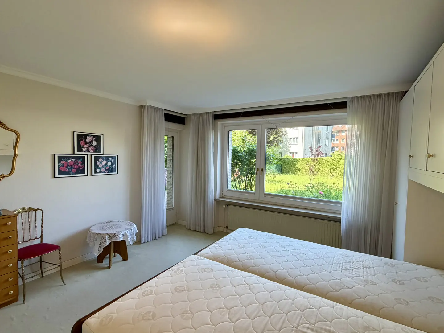Bright bedroom with two twin beds, a small table with a lace cloth, a vintage chair, and a large window overlooking a garden.