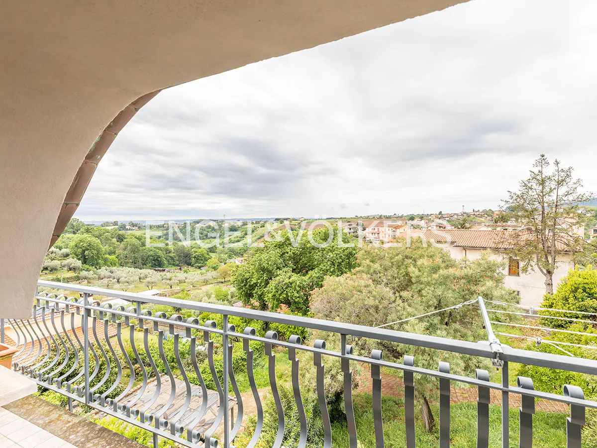 View from a balcony with gray metal railings overlooking green trees and houses under a cloudy sky.