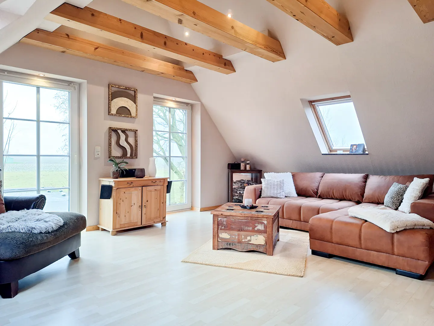 Attic living room with exposed wood beams, a brown leather sectional sofa, and a rustic wood coffee table on a light rug.