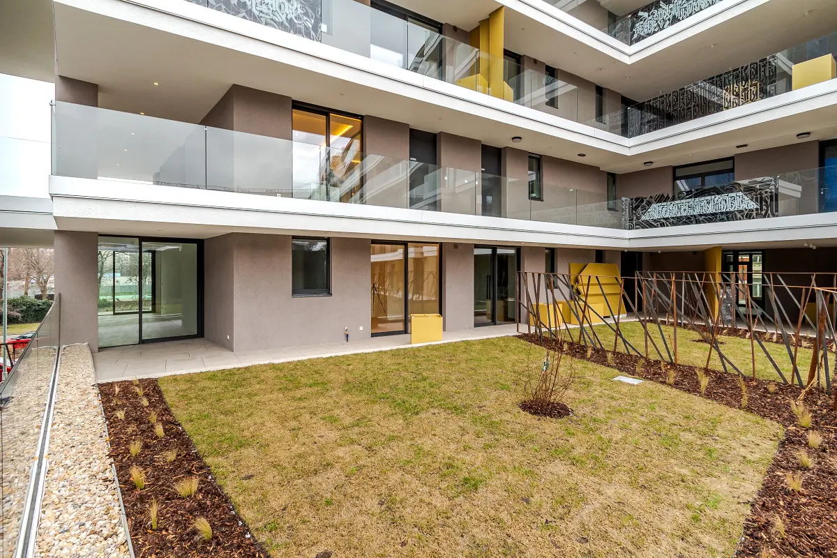 Apartment building courtyard with a lawn, mulch, and metal fence. Balconies with glass railings are visible.