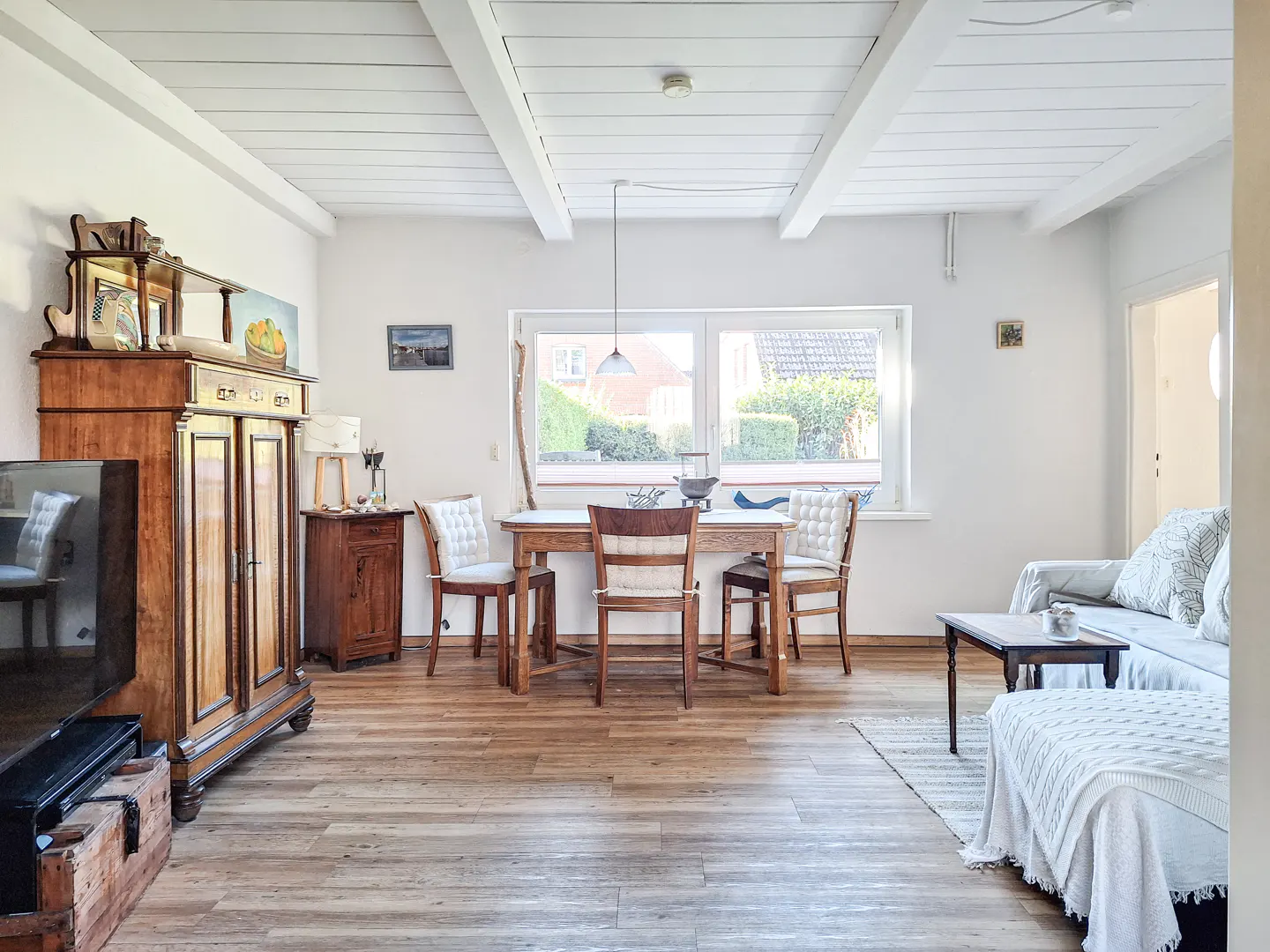 Bright living room with wood floors, white walls, and a beamed ceiling. A wooden dining table sits near a large window. A brown cabinet is on the left.