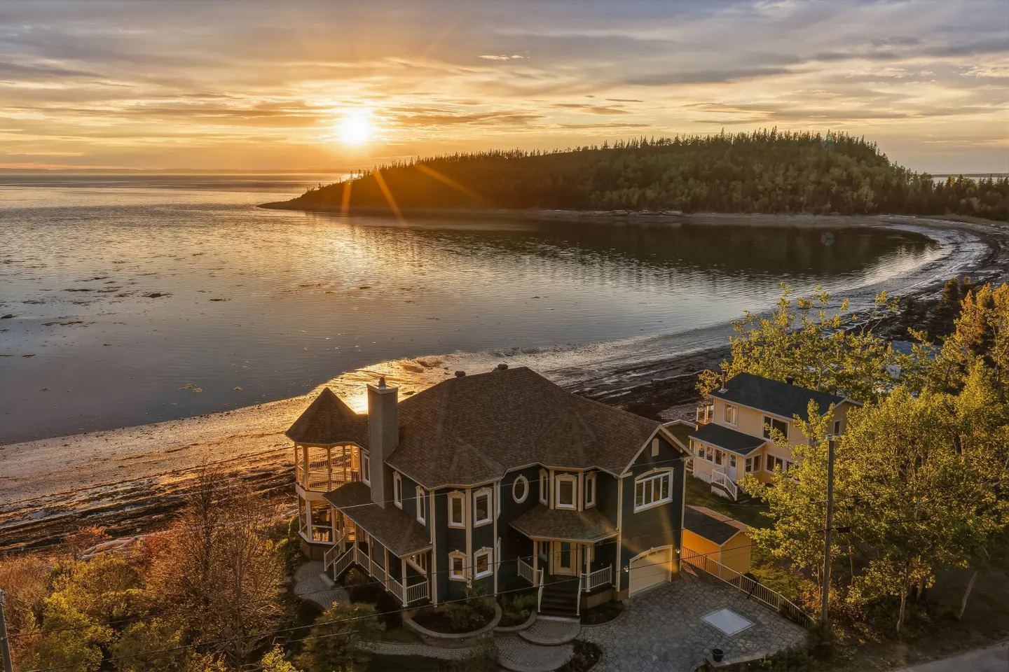 Aerial view of a dark green house on a beach at sunset, with a smaller yellow house nearby and a forested coastline.