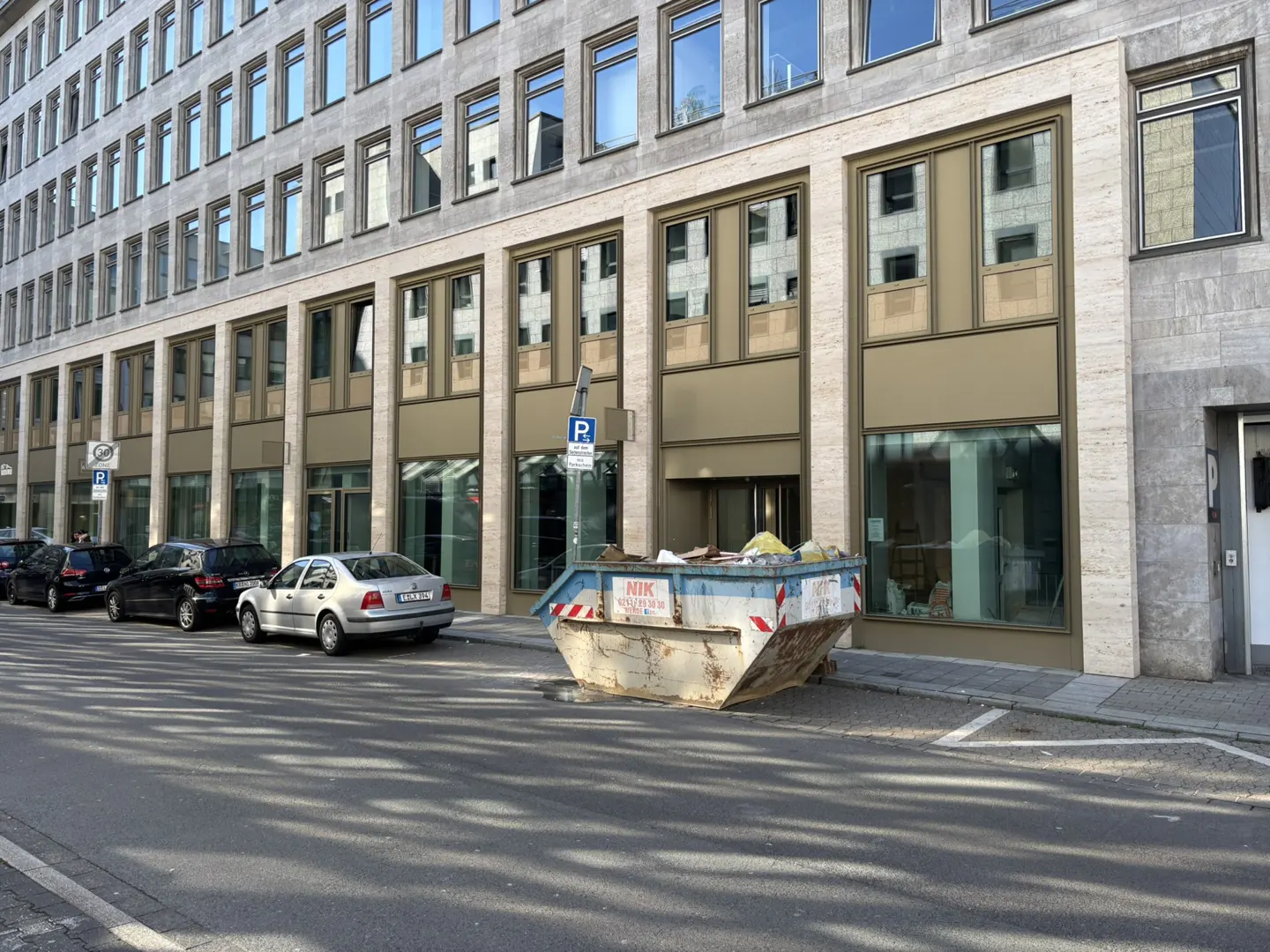 Street view of a gray building with many windows, parked cars, and a blue dumpster.