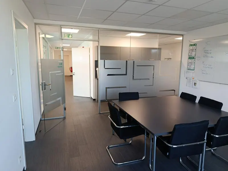 Office space with a large black conference table and chairs. Glass walls and doors provide light and visibility. A whiteboard is on the right.