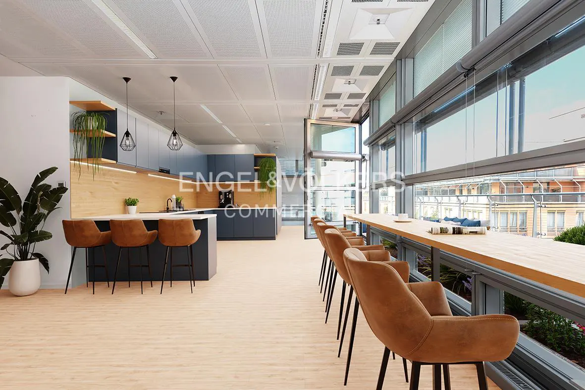 Open office space with a kitchen, bar stools, and a long table by the window. The kitchen has blue cabinets and wood accents. The floor is light wood.
