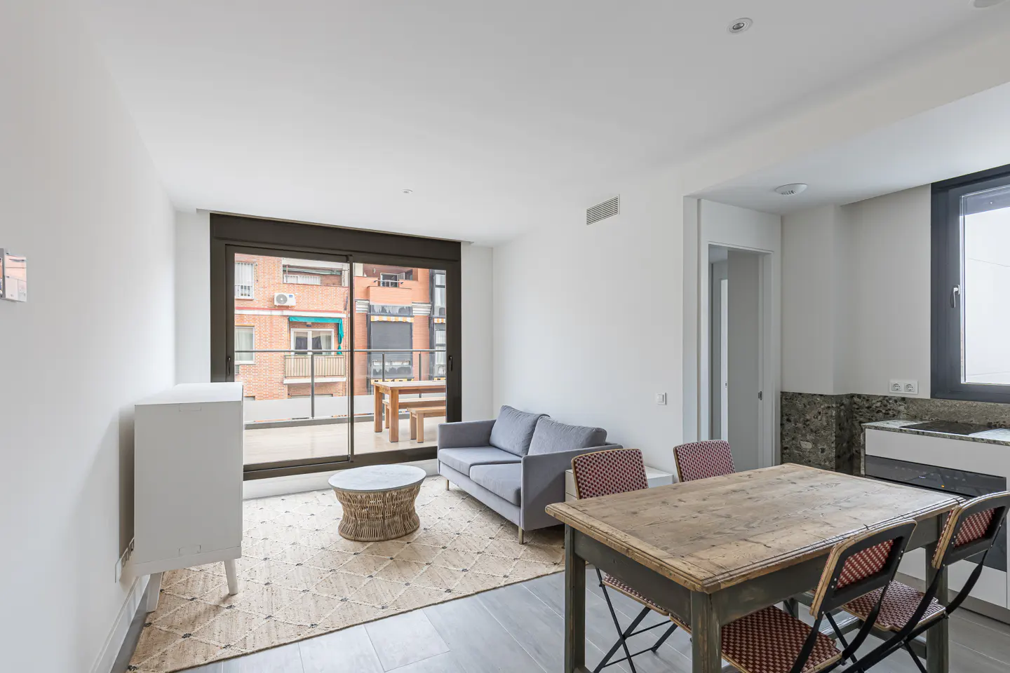 Bright living room with a gray sofa, woven rug, and rustic wood table with four chairs. Balcony doors open to a city view.