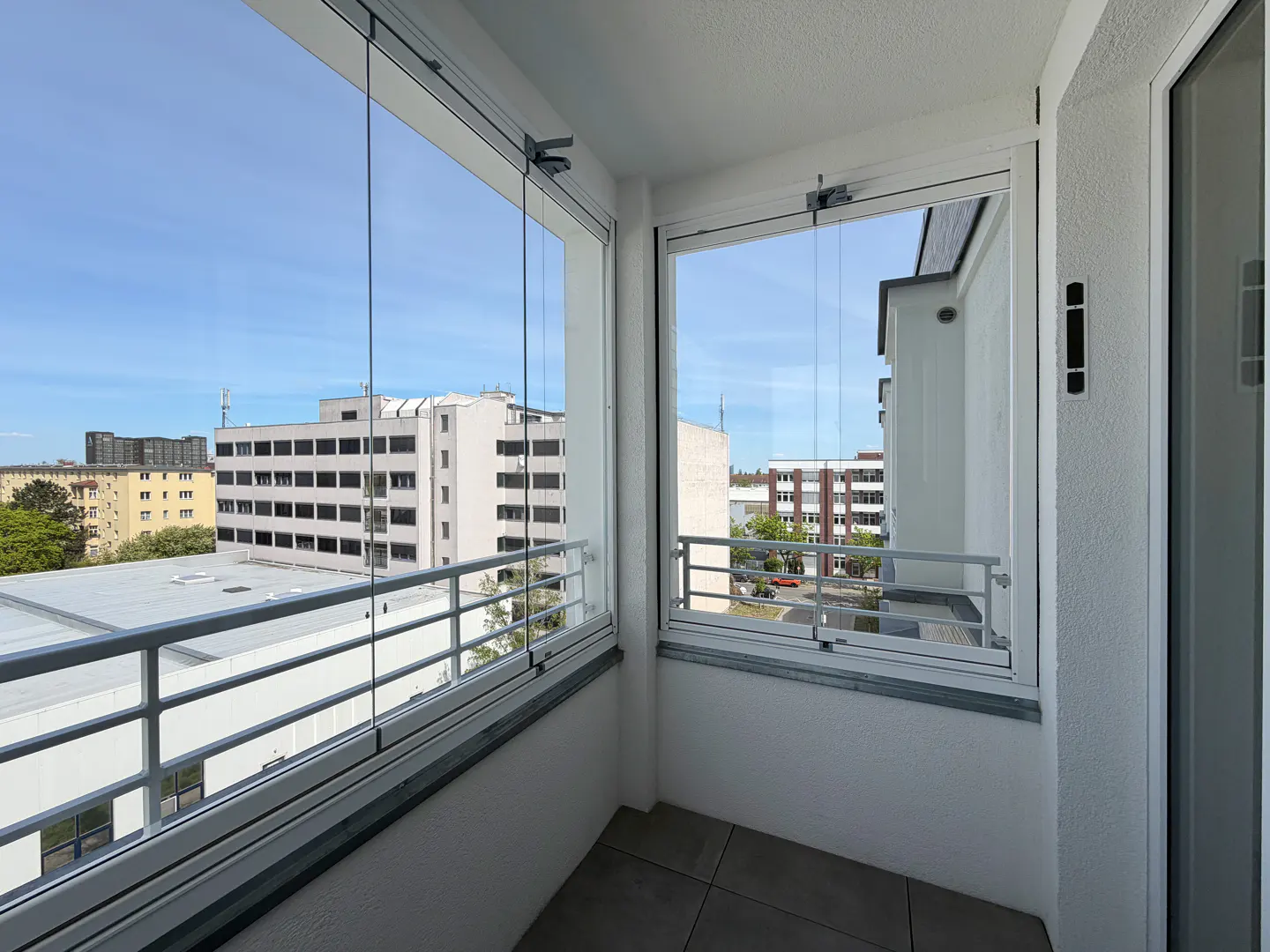 Balcony with retractable glass walls, white railings, and city views under a blue sky.