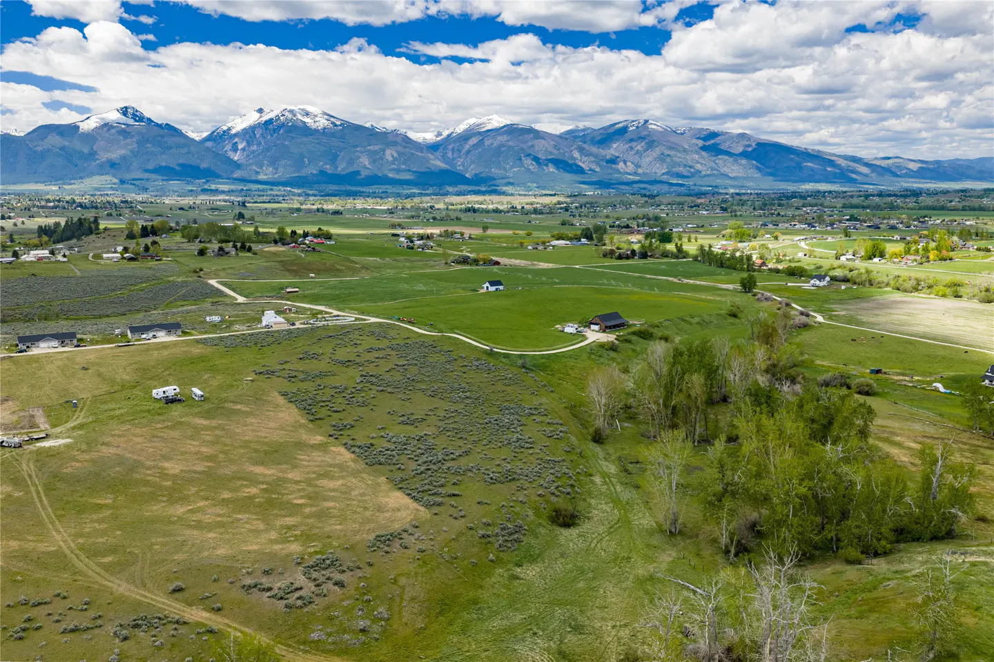 Aerial view of a green valley with houses, trees, and snow-capped mountains in the background under a blue sky with white clouds.