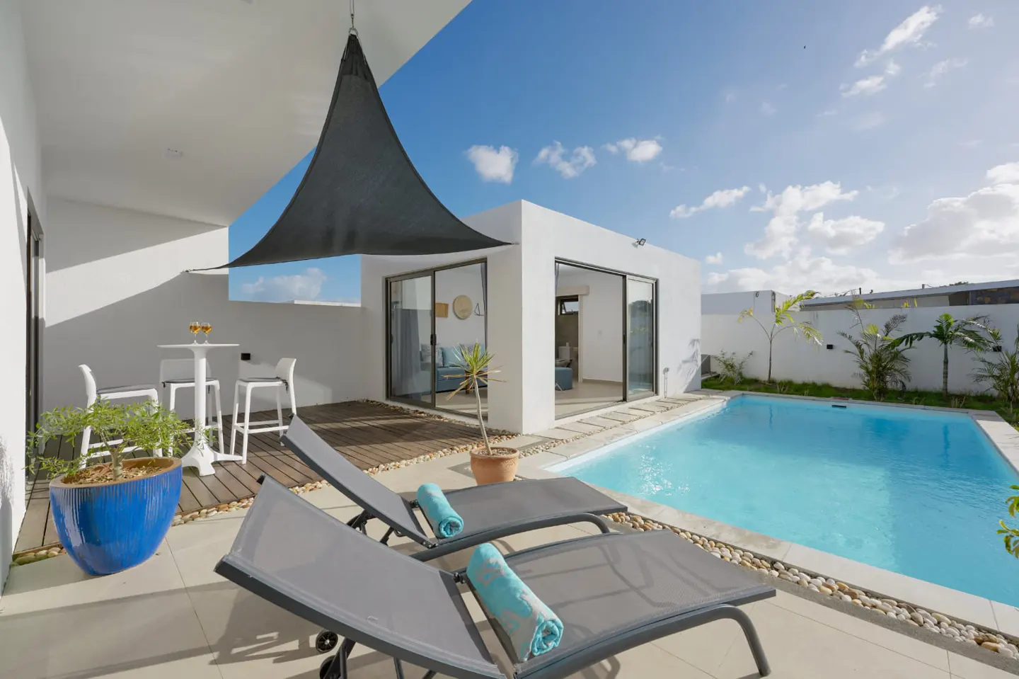 Modern villa patio with pool. Two gray lounge chairs with blue towels face the pool. White building with sliding glass doors. Blue sky.