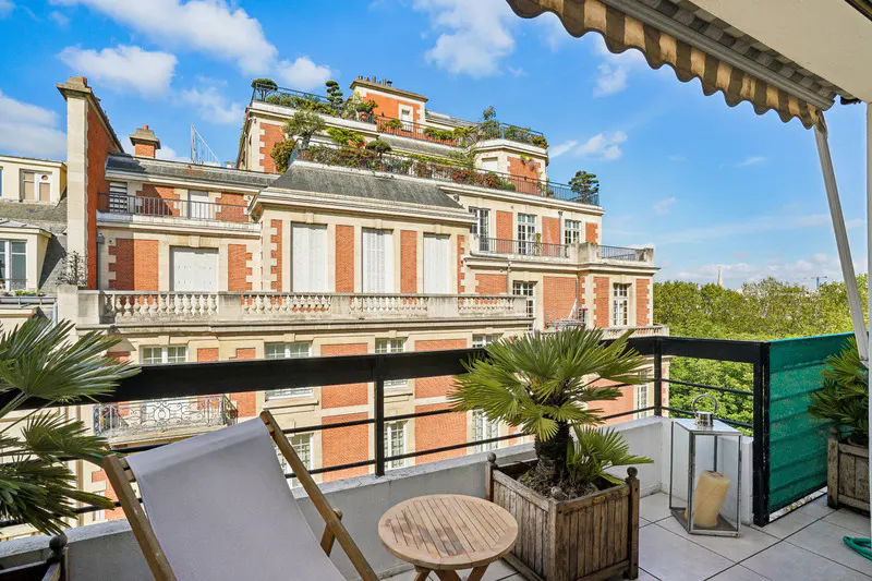 Balcony view of a red brick building with a white lounge chair, table, and potted palm tree.
