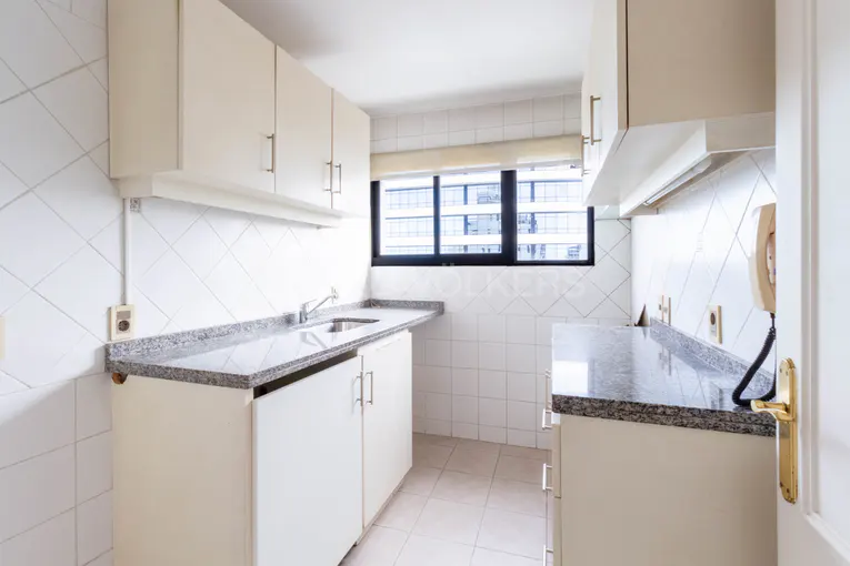 A bright kitchen with white cabinets, gray granite countertops, and white tiled walls. A window shows a city view.