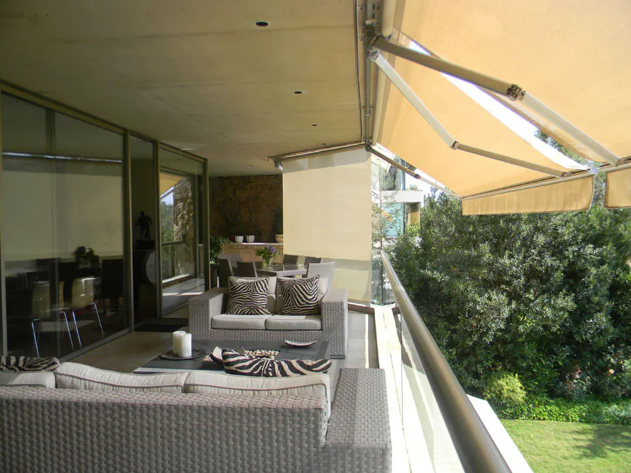 Outdoor patio with gray wicker furniture, zebra print pillows, and beige retractable awnings overlooking green trees.