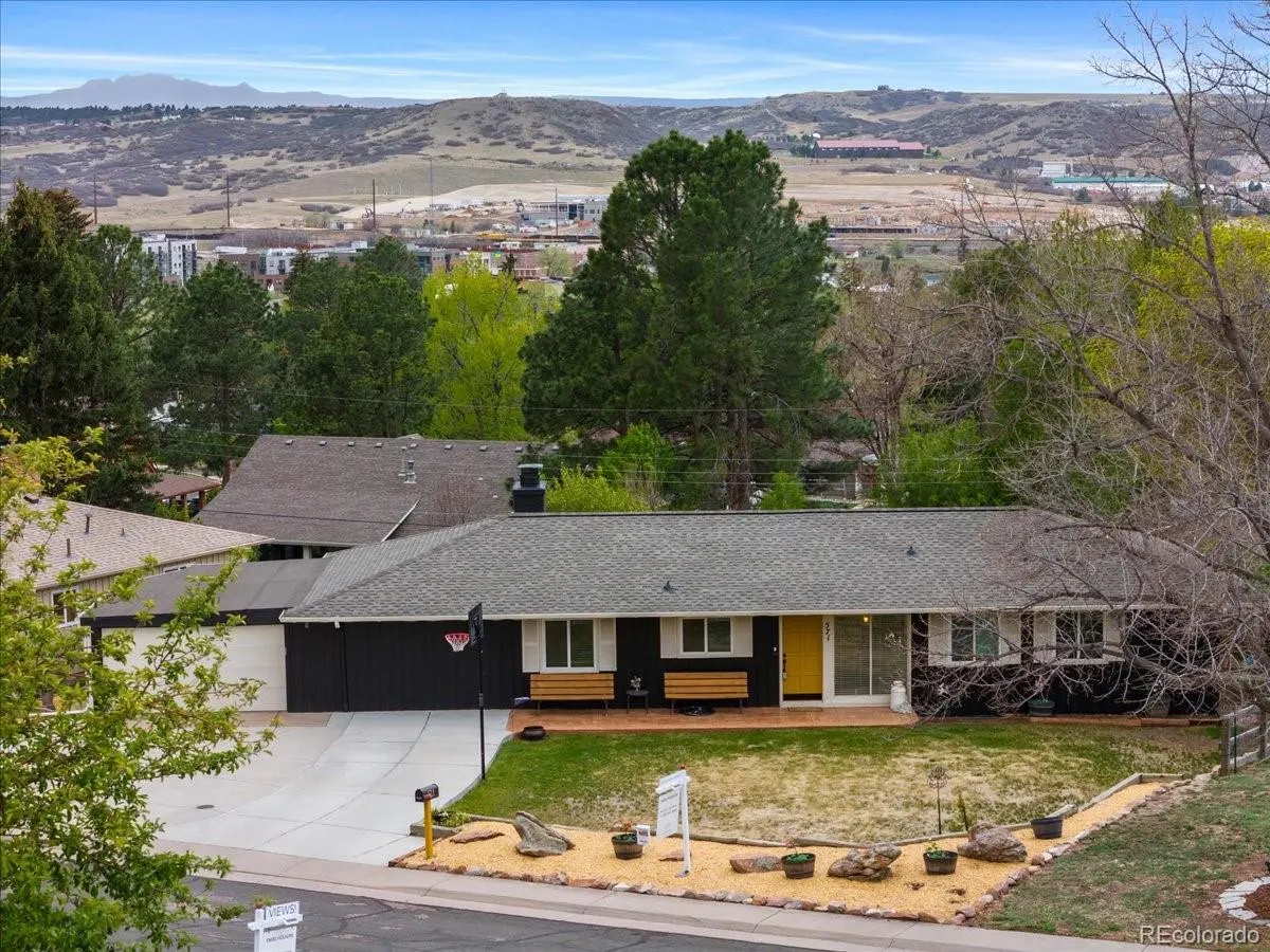 A single-story dark house with a yellow door, benches, and a basketball hoop in front of a mountain landscape.