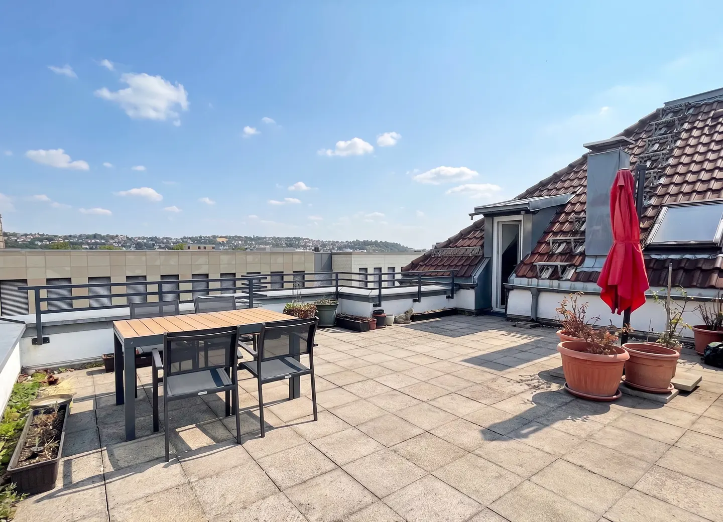 Rooftop patio with table, chairs, and potted plants. Red umbrella near a building with a red tile roof. Cityscape in the background.
