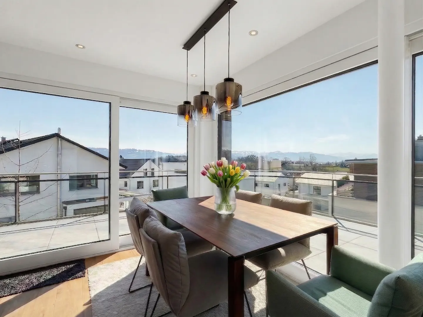 Bright dining room with a wood table, chairs, and tulips. Large windows offer a view of houses and mountains.