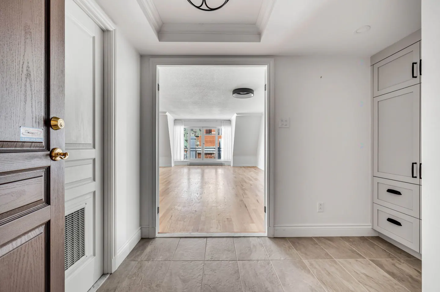 Open front door reveals a bright, white hallway with tile floor. A doorway leads to a room with hardwood floors and a window. White cabinets are on the right.