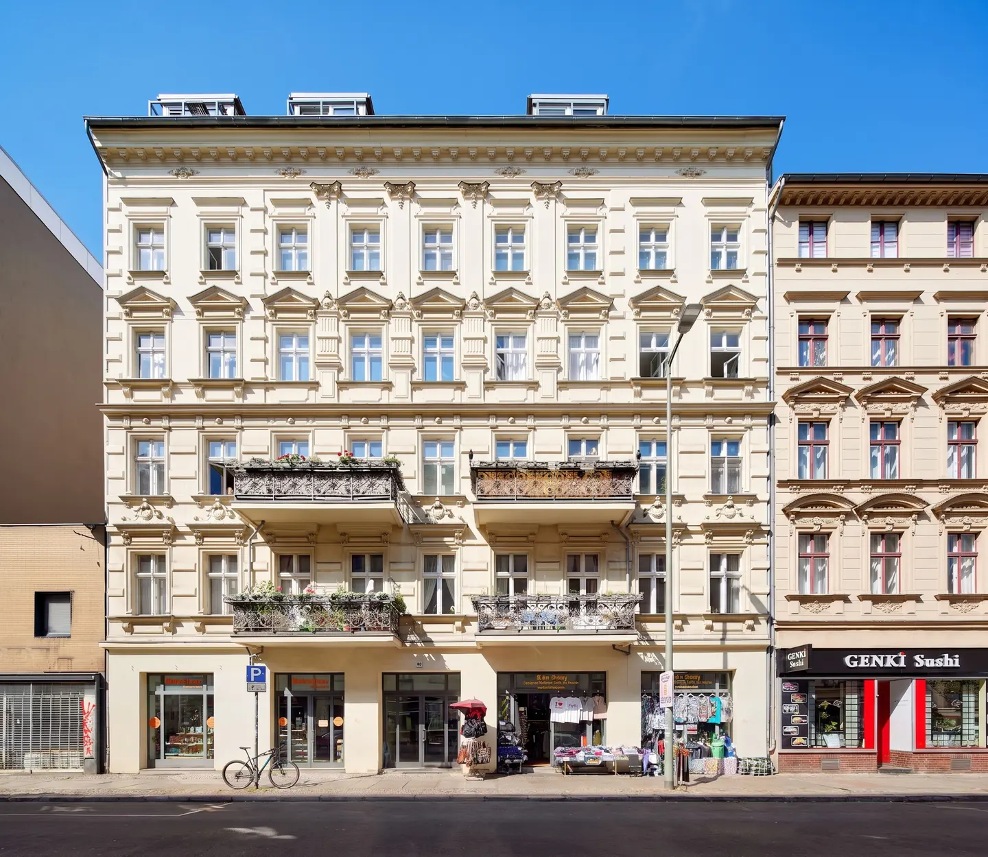 A five-story beige building with balconies and ornate window trim under a clear blue sky. Street-level shops are visible.