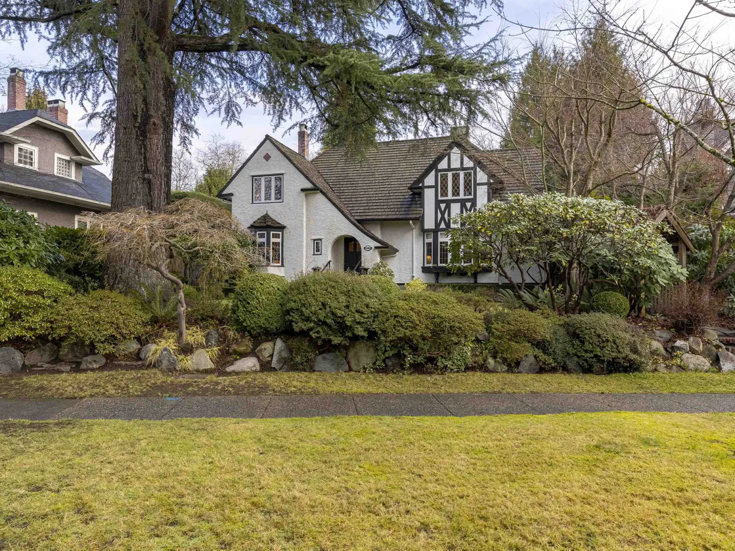 A white Tudor-style house with black trim sits on a landscaped lot with green grass and bushes.