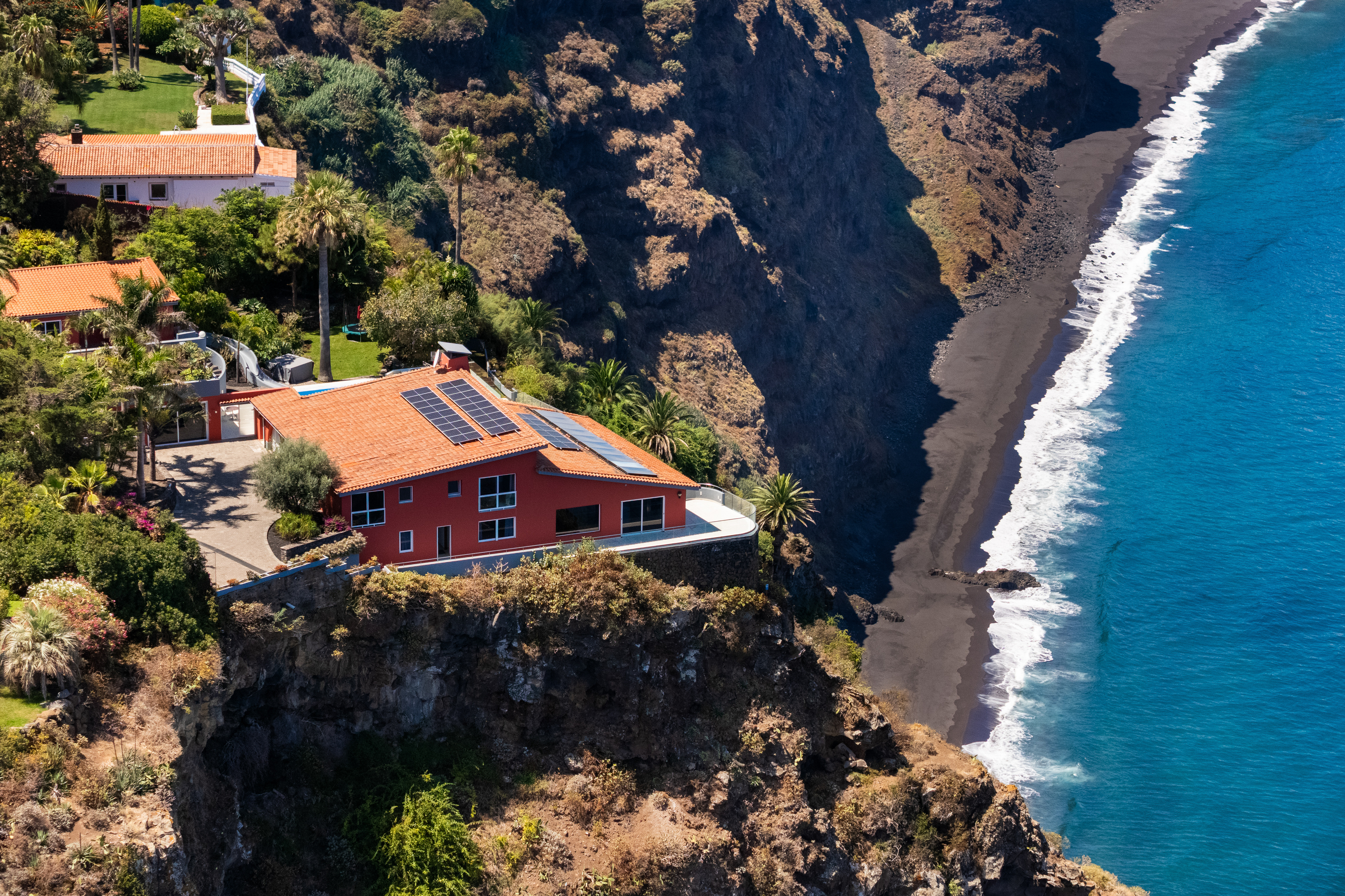 Aerial view of a red house with solar panels perched on a cliff above a black sand beach and the blue ocean. [AI-generated text]