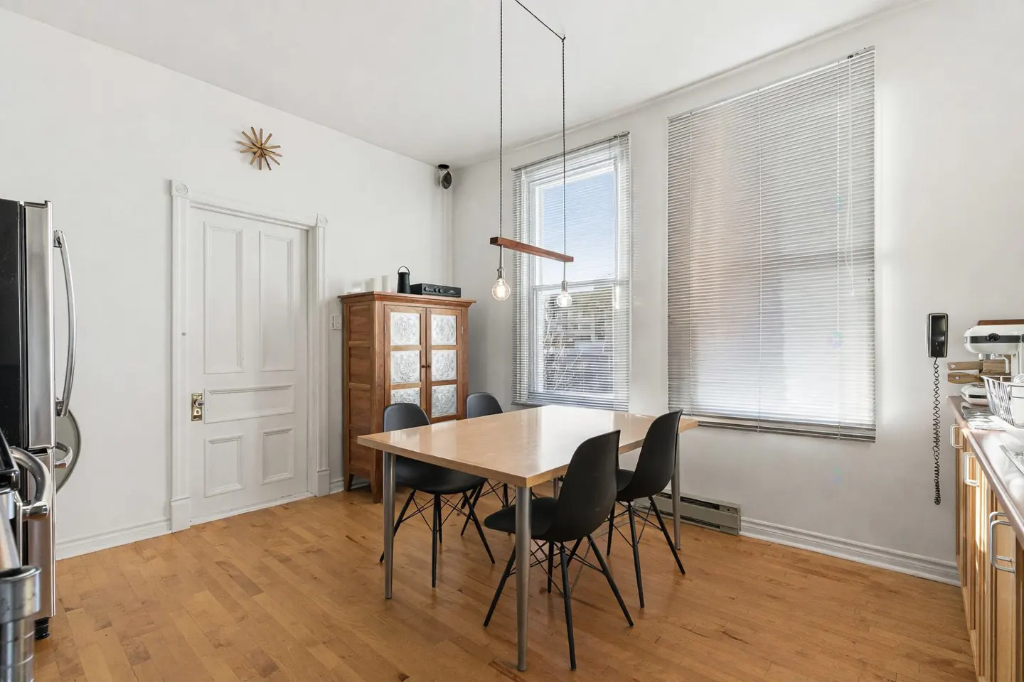 A bright dining area with a light wood table, four black chairs, and a modern light fixture. A wooden cabinet and white door are visible.