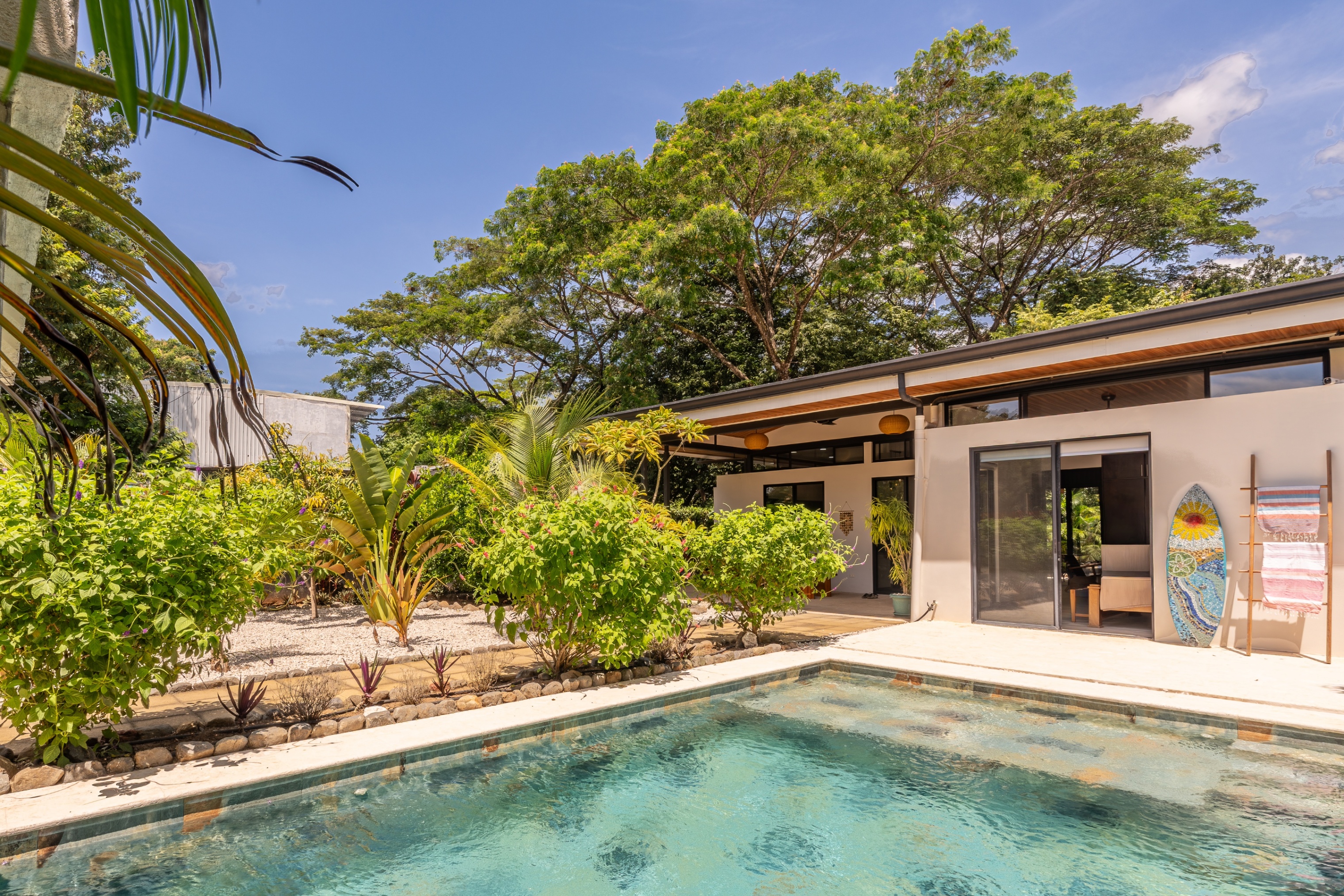 A modern home with a swimming pool, lush tropical landscaping, and a decorative surfboard leaning against the wall. [AI-generated text]