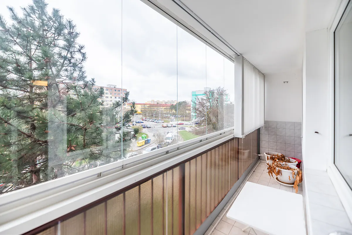Enclosed balcony with glass panels showing a city view. A small white table and dead plants in pots are on the tiled floor.