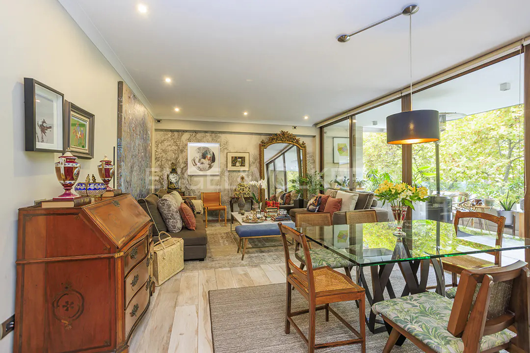 A bright living room with a glass table, wooden chairs, and a large window overlooking greenery.