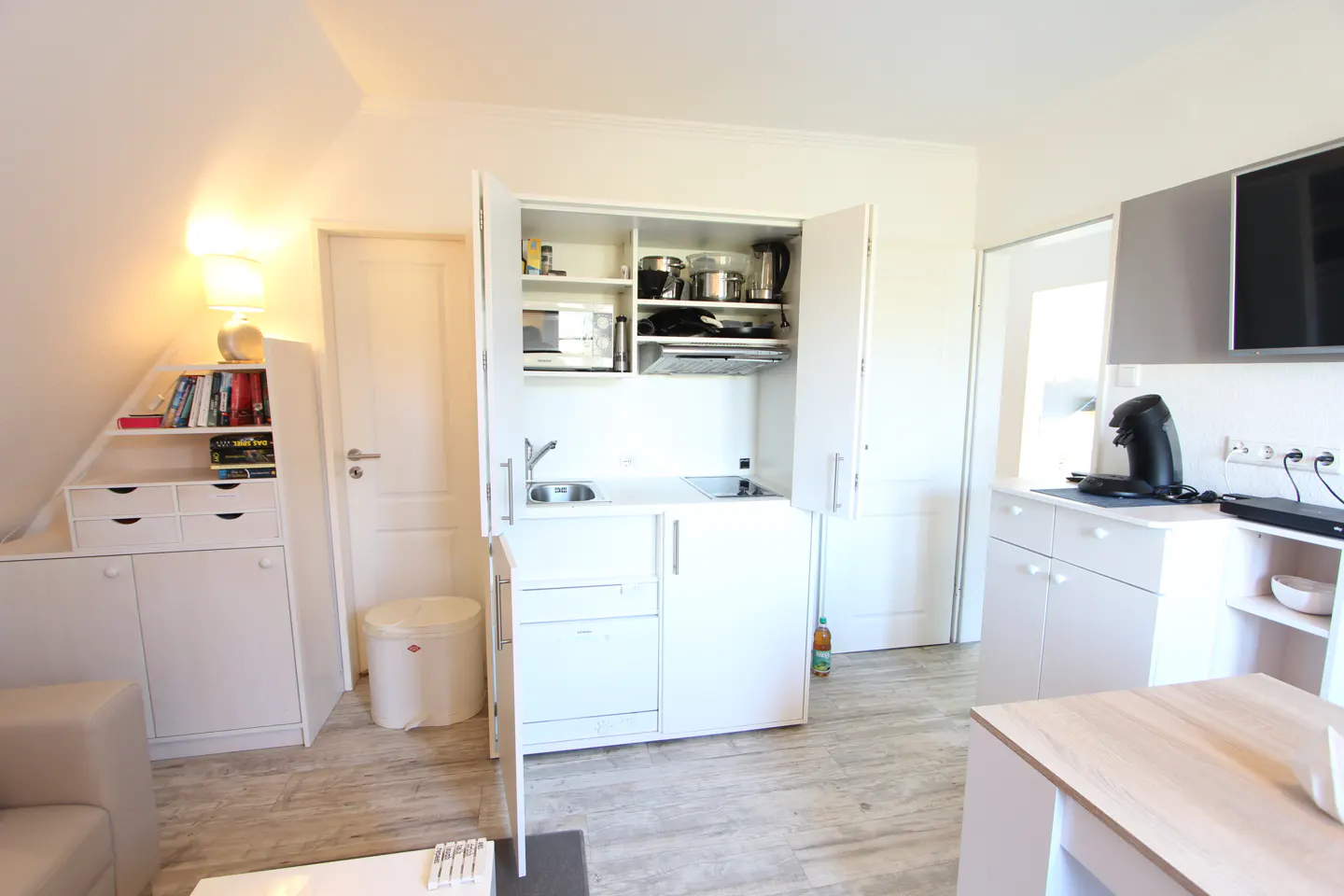 Bright, all-white studio apartment with a compact kitchen unit. The unit has open doors, revealing a sink, microwave, and cookware. A TV and coffee maker are visible in the background.