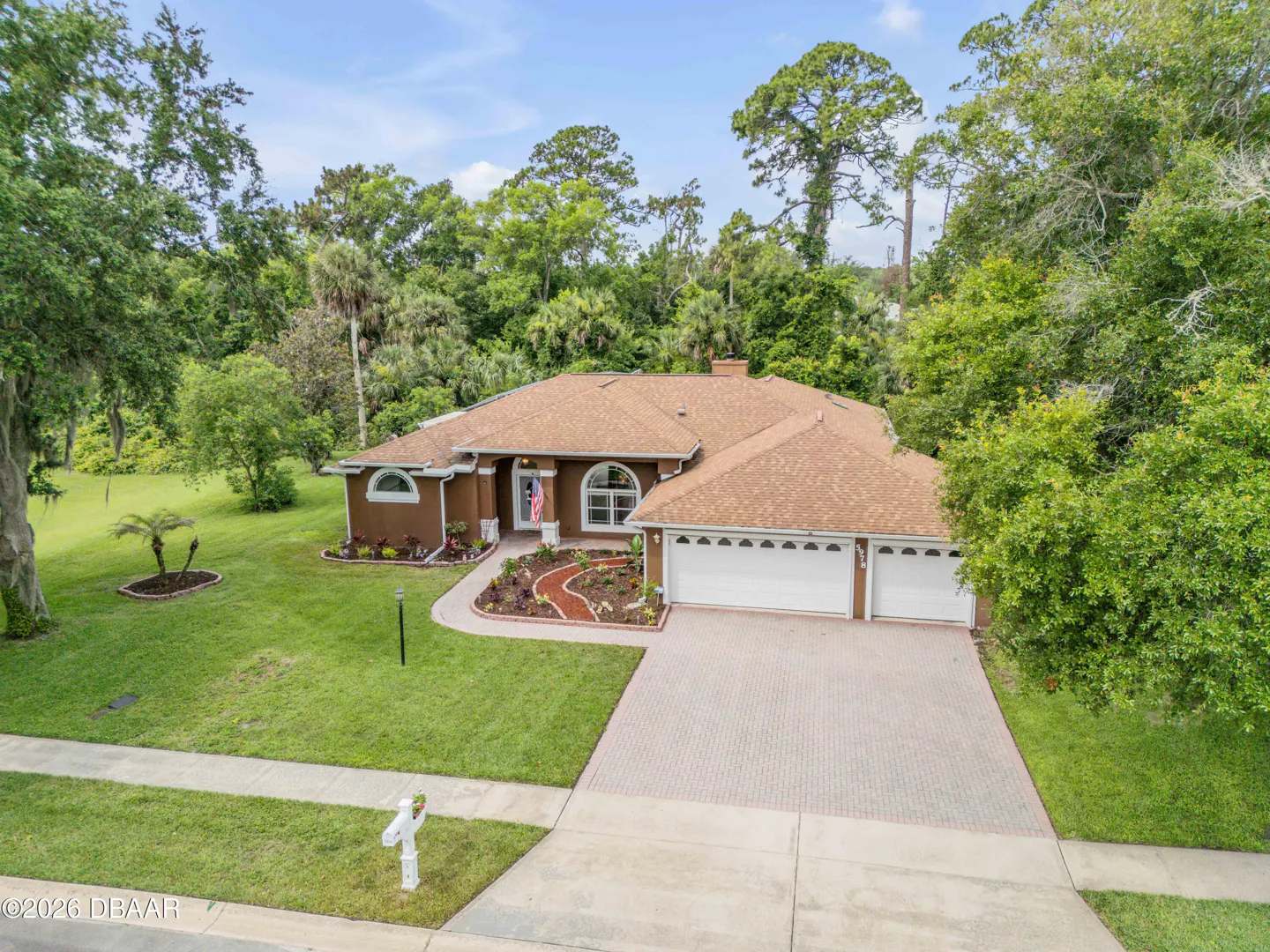 Single-story brown house with a tan shingle roof, white garage doors, and a paved driveway surrounded by lush green trees.